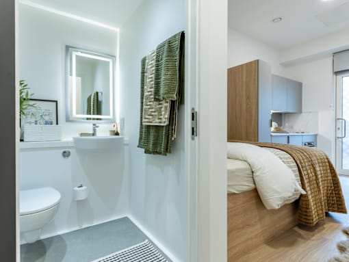 A bathroom with a toilet, sink, mirror, and a bedroom with a bed and a nightstand.