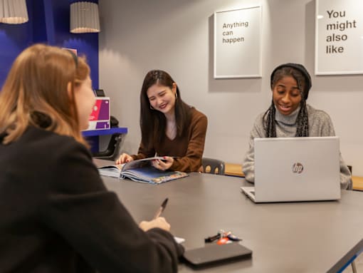 Three people are sitting at a table with a laptop, notebook, and pen.