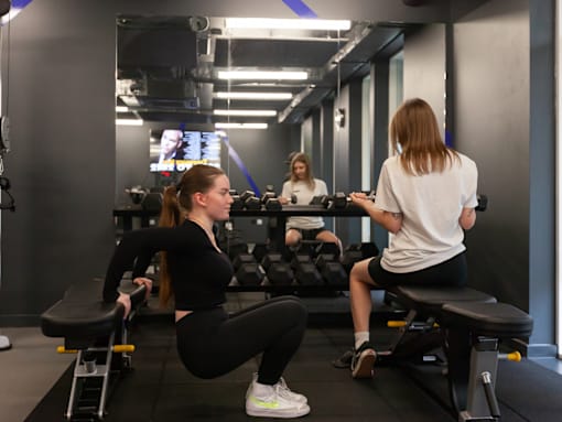 A woman is doing squats in a gym with another woman sitting on a bench in the background.
