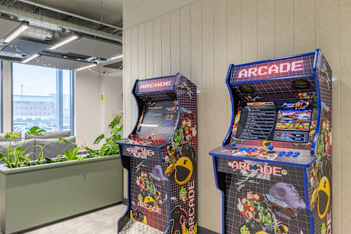 Two arcade machines are in a room with a window and plants.