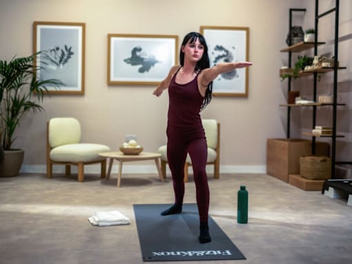A woman in a maroon top and leggings is standing on a yoga mat in a room with a chair, a table, and two framed pictures on the wall.