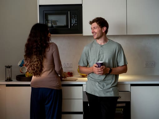 A man and a woman are standing in a kitchen, the man is holding a cup and they are both looking at a microwave.