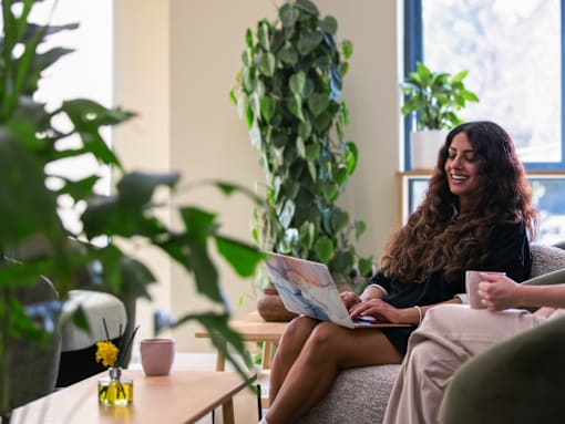 A woman is sitting on a couch with a laptop in a room with plants.