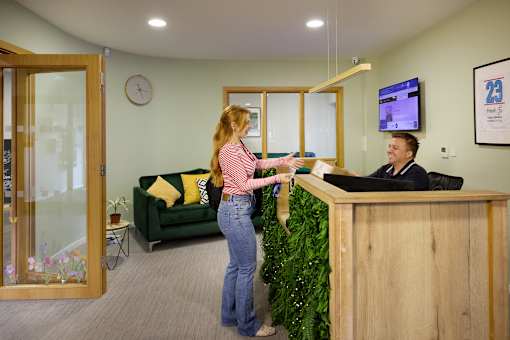 A woman in a pink top and blue jeans is handing a green plant to a man at a reception desk.