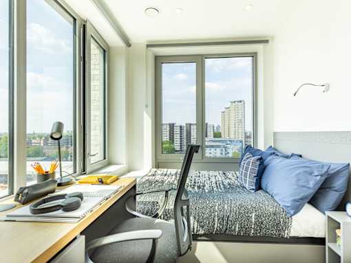 A bedroom with a bed, desk, and chair with a window overlooking a cityscape.