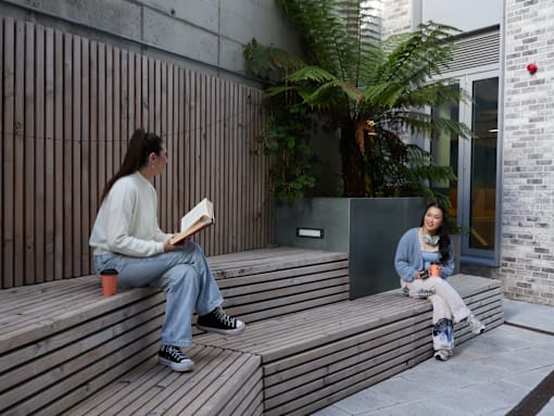 A woman is sitting on a wooden bench reading a book while another woman is sitting beside her on a skateboard.