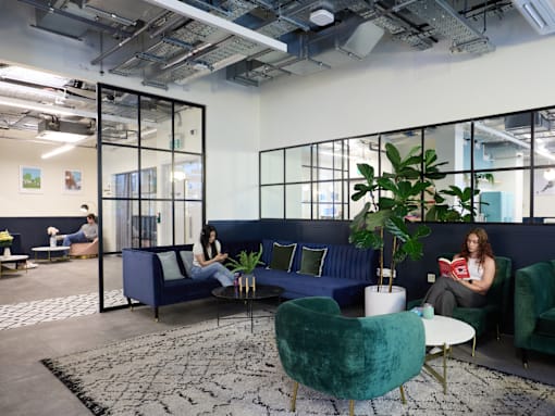 A woman is sitting on a blue couch reading a book in a modern office space.