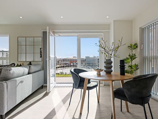 A modern living room with a grey sofa, a wooden table with two chairs, and a view of the city through the windows.