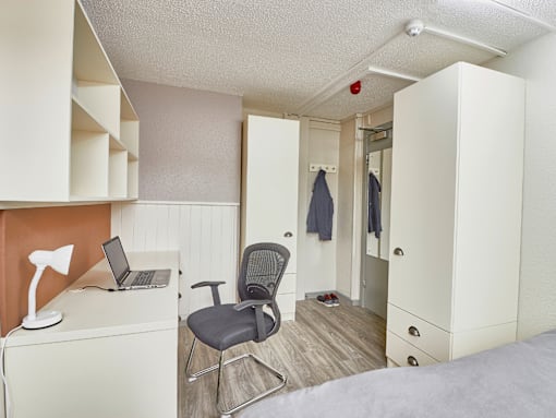 A room with a bed, a desk with a laptop and a chair.