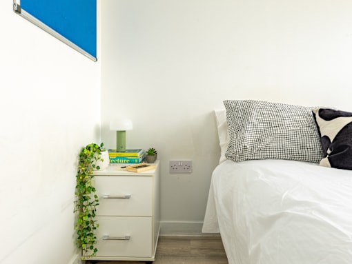 A white bed with a grey pillow and a white nightstand with a green lamp and a book on it.