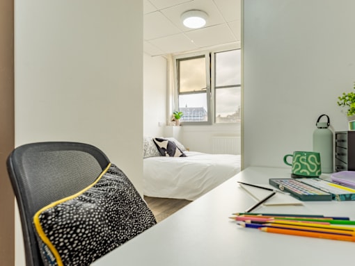 A black chair with a yellow stripe is in front of a white desk with a white bed in the background.