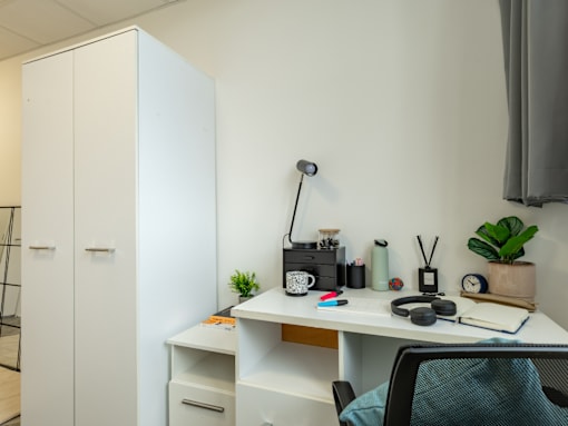 A white desk with a laptop, a cup, a camera, a notebook, a pen, a pair of headphones, a plant, and a black chair.