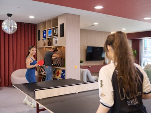 Two women playing ping pong in a room with red curtains.