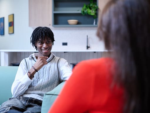 A man with dreadlocks is sitting on a green couch and smiling at a woman in a red top.
