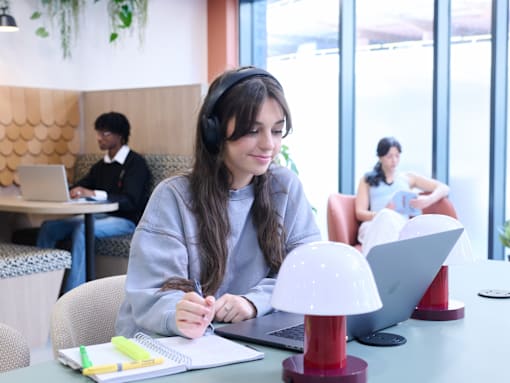 A person is writing in a notebook with a laptop and a lamp on the table.