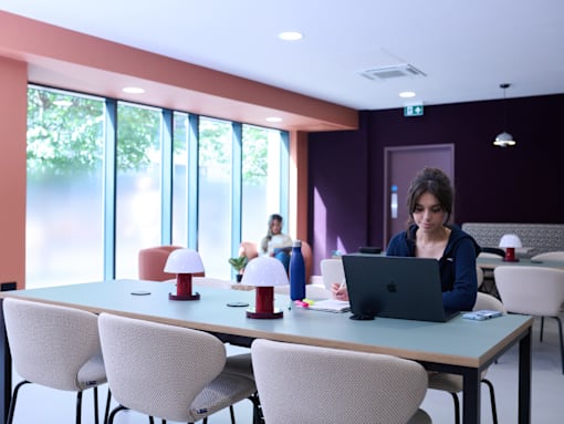 A woman is working on her laptop in a room with a table, chairs, and a window.