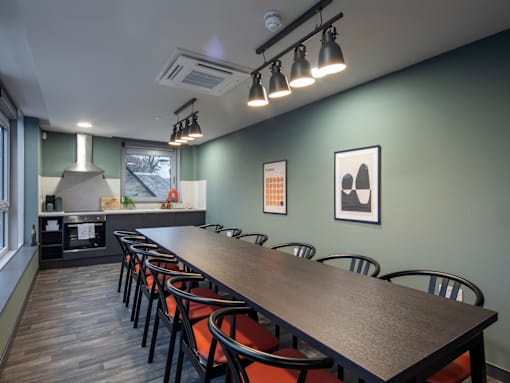 A long wooden table is surrounded by chairs in a room with a kitchen area in the background.