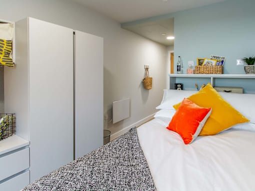 Bedroom with a bed and wardrobe. The bed is decorated with a grey throw, white bedding and orange/yellow pillows.