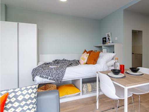 Side-view angle of a bed, with white bedding, a grey throw and yellow/orange pillows. Includes view of under-bed storage and a small dining table and chairs.