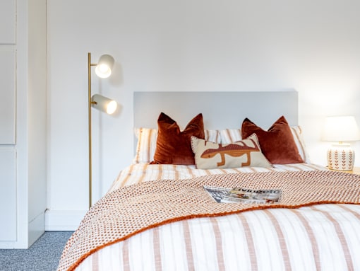 A bed with a white headboard and orange pillows with a white lamp on the side.