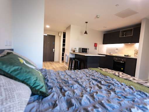A bedroom with a bed and a kitchen area in the background.