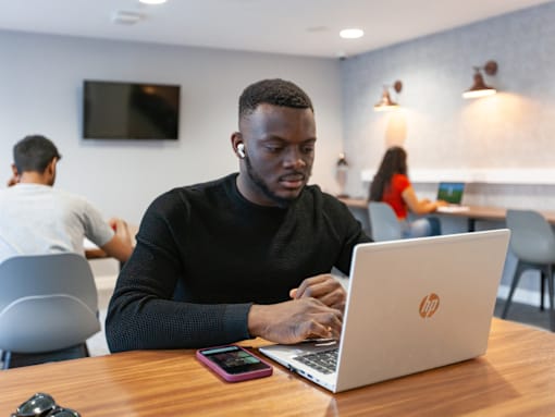 A man in a black shirt is using a laptop in a room with other people in the background.