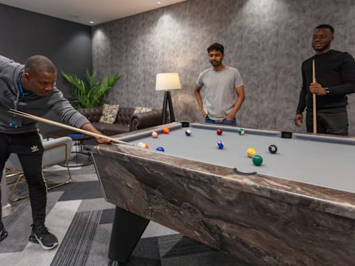 Three men playing pool in a room with a grey couch and a plant.