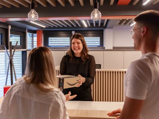 A woman in a black dress is holding a box with the Amazon logo on it and talking to two people.