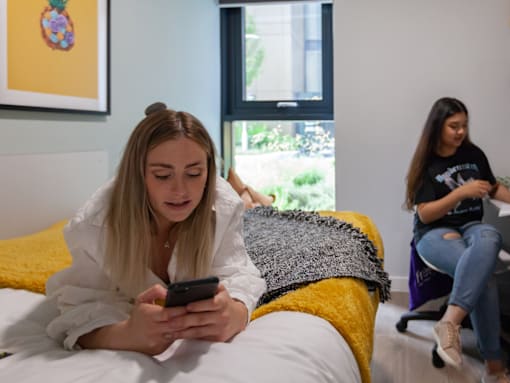 Two girls are sitting in a room, one is on a bed and the other is on a chair.