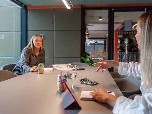 Two women having a conversation over a table with a can, glass of water, and a calculator on it.