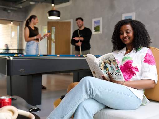 A woman is sitting on a couch reading a magazine while two other people play pool in the background.