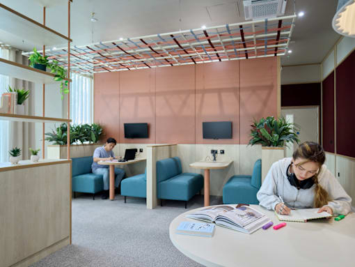 A woman is studying at a desk in a room with a pink wall and a wooden ceiling.