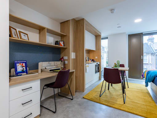 A room with a desk, chairs, and a kitchen area.