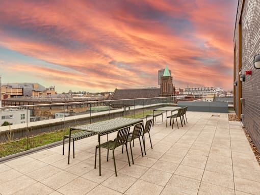 A rooftop patio with a table and chairs overlooking a cityscape at sunset.