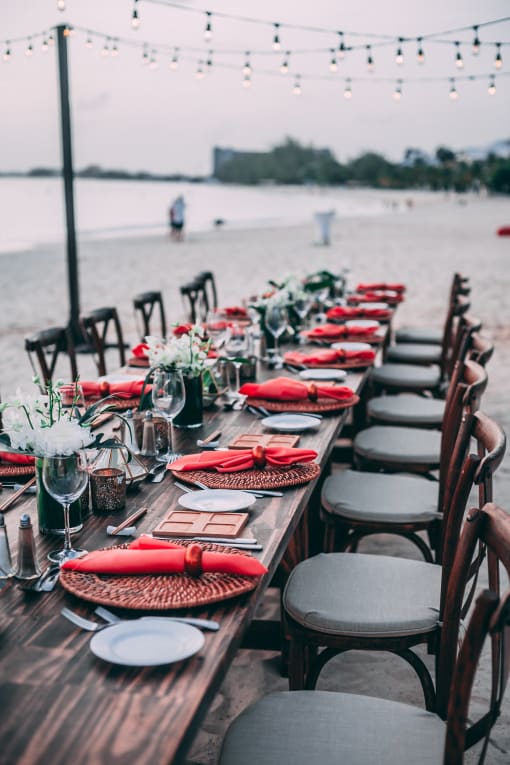 a long wooden table with red napkins and chairs at the beach