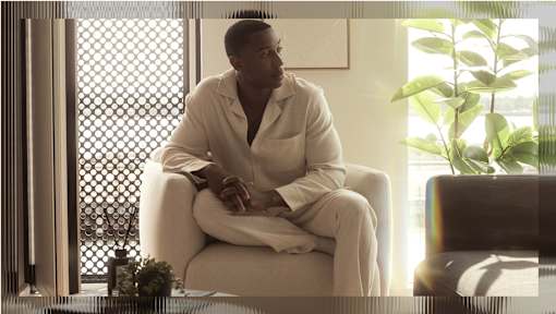 A man in a white robe sits on a chair by a window.