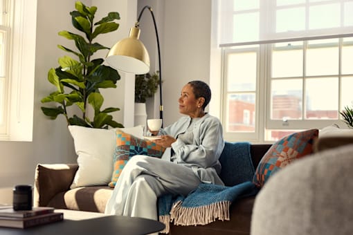 A woman in pajamas sits on a couch with a cup of coffee.