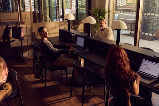 Three people are working on laptops in a room with large windows.