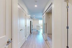 Modern, bright apartment hallway with wood flooring, white walls, and multiple doors. An open kitchen with stainless steel appliances is visible ahead.