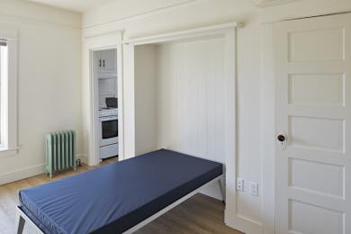 A bed with a blue cover sits in a room with white walls and a white door.