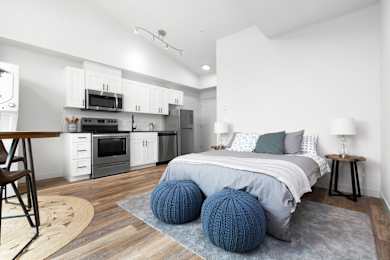 Floor Plan  A modern bedroom with a bed, two blue knitted poufs, and a kitchen area with white cabinets and appliances.