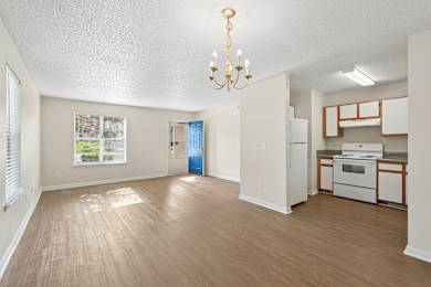 A well-lit, empty room with a chandelier and a kitchenette in the background.
