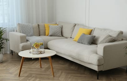 A grey couch with a yellow pillow and a small table with a vase of flowers on it.