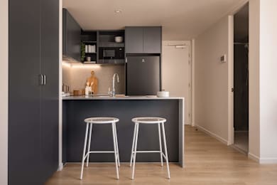 A kitchen with a black fridge and cabinets, a wooden cutting board, and two white bar stools.