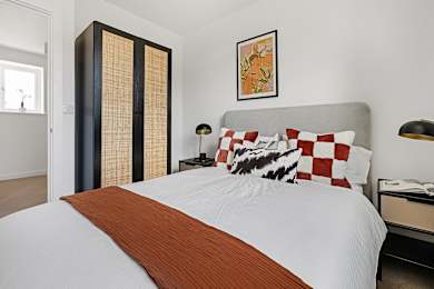 Floor Plan  A bed with a white comforter and a brown throw sits in a room with a tall wardrobe and a lamp.