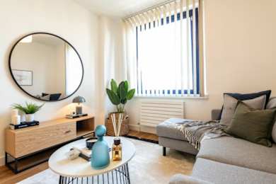 A living room with a grey couch, a round mirror, a wooden cabinet, a table with a blue vase, and a window with blinds at The Keel Apartments, Liverpool