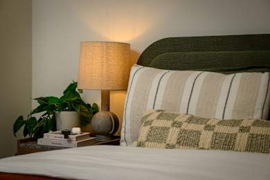 A lamp with a beige shade is on a bedside table next to a potted plant.