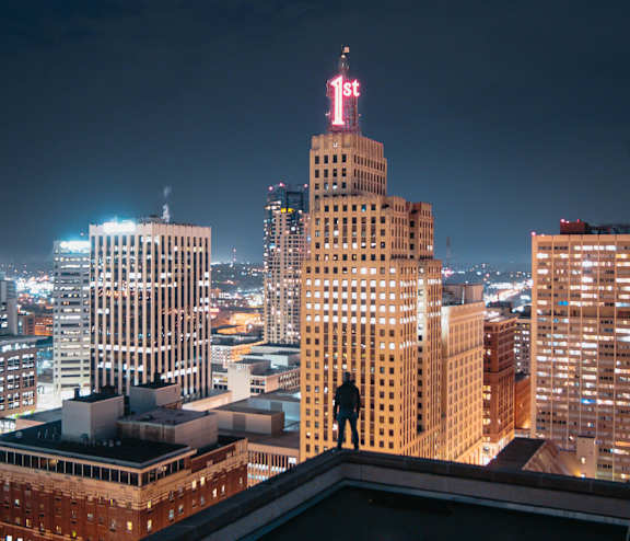 A man stands on the roof of a building looking at the city skyline at night.