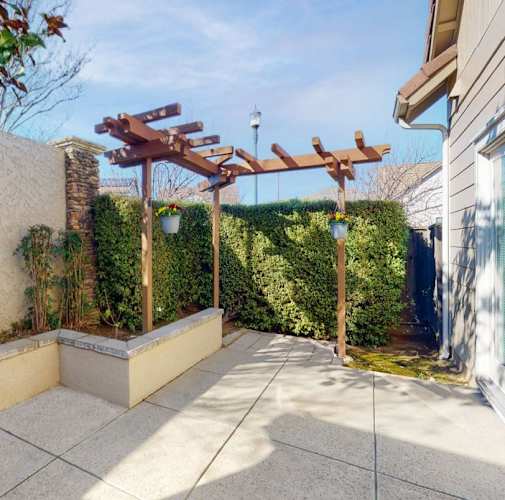 A wooden pergola is in the middle of a patio with a wall of green hedges.