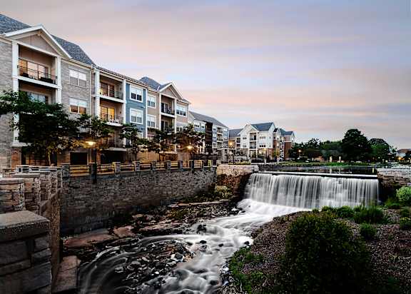 A waterfall flows into a river in front of apartment buildings.